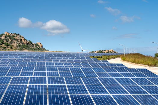 Expansive solar farm with rows of panels under clear blue sky and hills in the background.